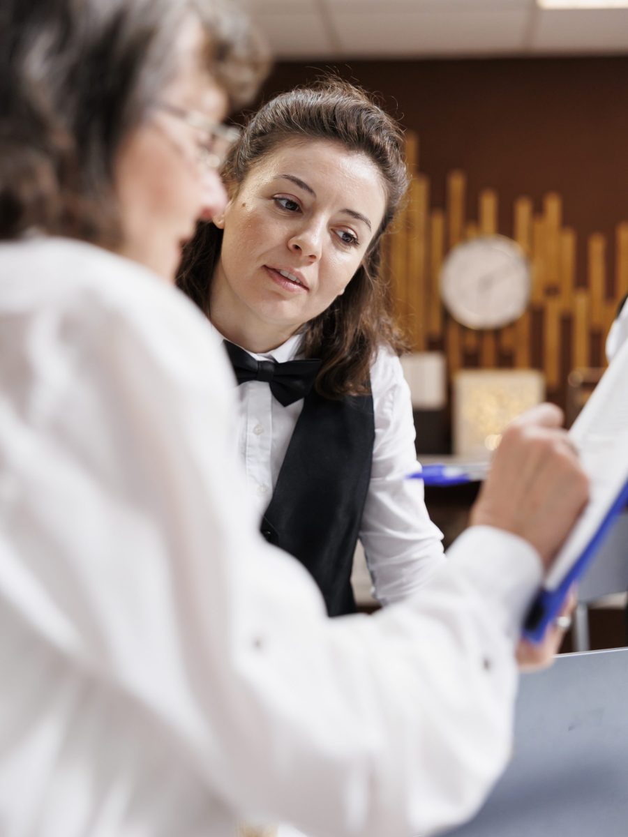 At hotel foyer, female concierge serves retired elderly woman, ensuring flawless check-in process. As she signs reservation paperwork, receptionist delivers polite assistance to old lady traveler.